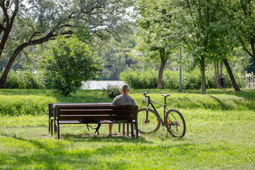 Elderly sports gray haired man resting on bench by the lake after riding bicycle. Beautiful natural landscape. Active lifestyle and sport activities in senior age concept