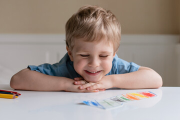 Cute little boy with blond hair and blue T-shirt draws colored pencils at home. Draws at the white table. Boy smiles.