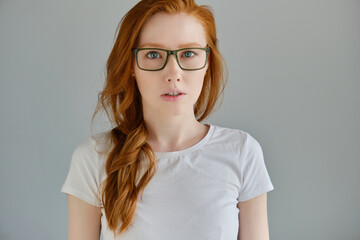 Headshot. Red-haired girl in a white T-shirt and glasses stands on a gray background and looks into the frame.