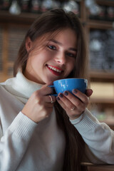 Young smiling girl is holding a cup of coffee. Woman with long hair is looking in the lens.
