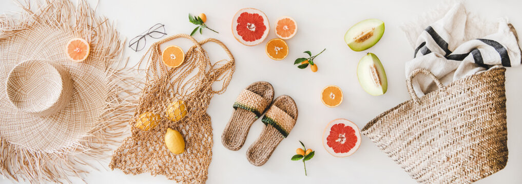 Summer mood layout. Flat-lay of summer natural sandals, straw sunhat, beach rafia and net bag, striped beach towel, sunglasses and cut fresh fruits over white plain background, top view