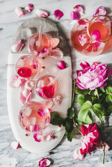 Summer refreshing cold beverage drink. Flat-lay of rose lemonade with ice in glasses and jug and fresh rose flower petals on oval serving board over grey marble table background, top view