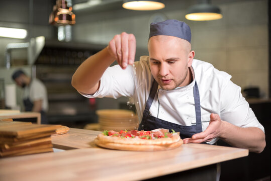 Food concept. Preparing traditional italian pizza. Young smiling chef in white uniform and gray hat decorate ready dish in interior of modern restaurant kitchen. Ready to eat