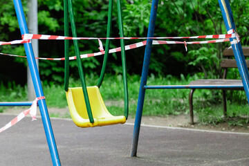 Fototapeta premium Yellow children's swing on a sports playground wrapped with red barrier tape. Prohibition of outdoor walks, prevention of the coronavirus influenza virus covid-19. infection. selective focus