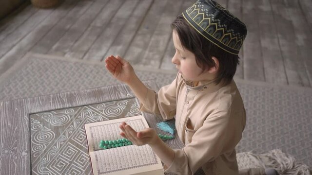 Adorable cute little boy in prayer hat and arabic clothes with rosary beads reading holy Koran book praying to Allah, prophet Muhammad holy spirit religion symbol concept inside eastern interior