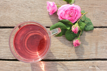 Tea rose herbal tea in a glass cup with pink buds and flowers on a rustic wooden table. Summer tea party outdoors
