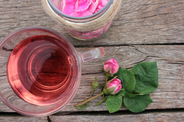 Tea rose herbal tea in a glass cup with pink buds and flowers on a rustic wooden table. Summer tea party outdoors
