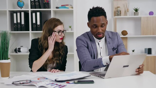 Purposeful considerate experienced young businesswoman and her black-skinned partner working together with documents and information from laptop in the office,4k