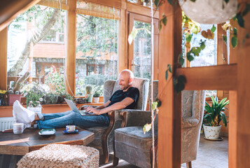 Man dressed black T-shirt and jeans sitting in a comfortable armchair, using a modern slim laptop...