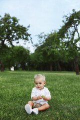 Little baby boy in white shirt and shorts sitting alone on the grass in summer day. Small kid 1-year-old in posing outdoors.