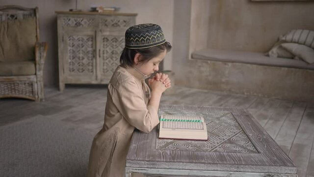 Little muslim boy in prayer hat and arabic clothes with rosary beads reading holy Quran book praying to Allah, prophet Muhammad holy spirit religion symbol concept inside eastern interior