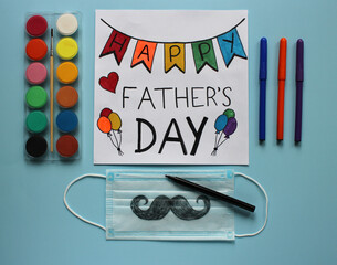 Greeting card Happy Father’s Day and protective face mask with a mustache drawn by a child as a gift for dad. Blue background. Concept of Father’s day in the context of coronavirus pandemic