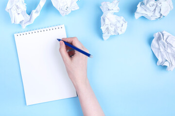 Woman writes in notebook around crumpled paper, blue background. Concept of beginning to draw up plan, discarding bad ideas. Flat lay. Copy space