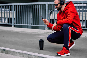 Young handsome sportsman crouching, resting and using smart phone after long running in city.
