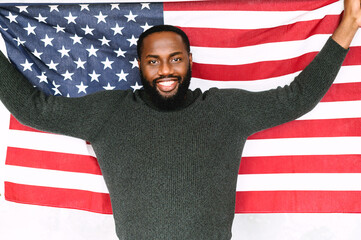 Young handsome smiling black guy stands with hands up and holding US flag behind. Studio shot