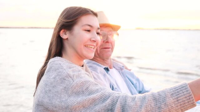 Make selfie or video call by happy talking young granddaughter kissing hugging old smiling grandfather near water at sunset. Modern lifestyle, enjoy family leisure using mobile device together outdoor