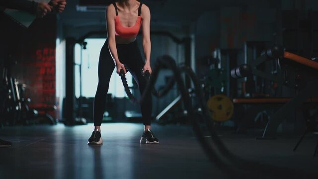athletic female exercising ropes in modern gym with personal trainer