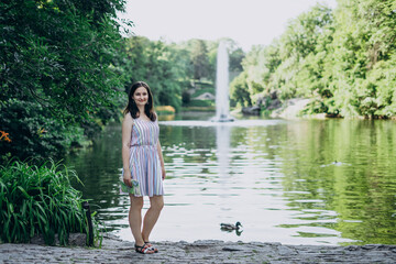 Uman, Ukraine. Smiling girl on a background of high fountain and lake. Girl in a summer dress on a background of a beautiful tall fountain. Lake in the park and splashing fountain.