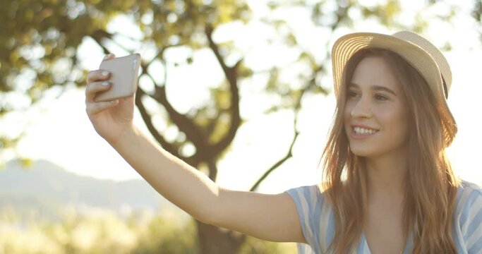 Beautiful Caucasian young girl with dark hair and in hat smiling and posing to smartphone camera on sunny day at nature. Pretty cheerful brunette female taking photos with phone in summer.