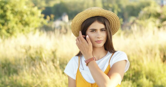 Portrait shot of beautiful young brunette Caucasian woman in hat tanding in field and looking at camera. Charming girl with dark hair at nature place in summer. Outdoor.