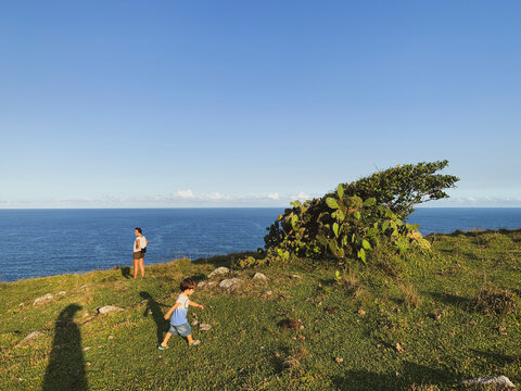 Woman and little boy near cactus and tree on green hill close to the sea at the Atlantic ocean coast. Panoramic image on a sunny day with blue sky and no clouds.