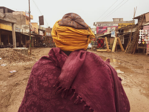 Person from behind wearing turban scarf and poncho walking on mud dirt street after the rain in poor neighborhood.