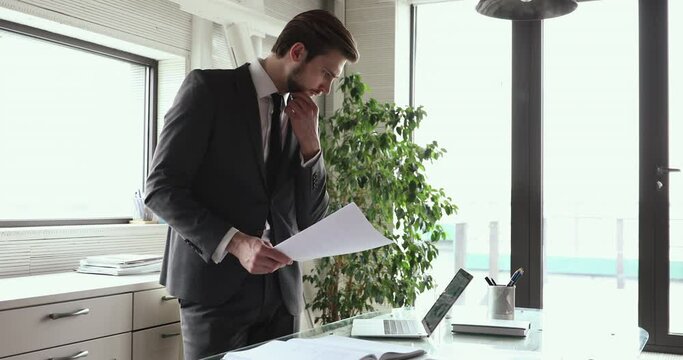 Thoughtful serious male ceo wearing suit concentrating planning strategy working on laptop computer doing paperwork standing at workplace. Focused businessman thinking of business planning in office.