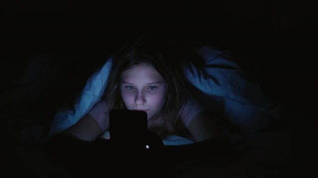 Youth addiction. Online night. Teen girl hiding with phone under blanket in bed.