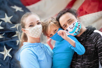 Family members embracing each other, smiling in the camera wearing cloth face masks with the US flag on the background. It's recommended to cover faces during the world coronavirus covid-19 pandemic.