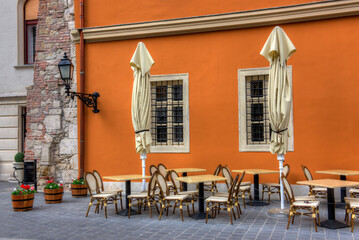 A closed up restaurant with locked tables, chairs and umbrellas with an orange painted outside wall