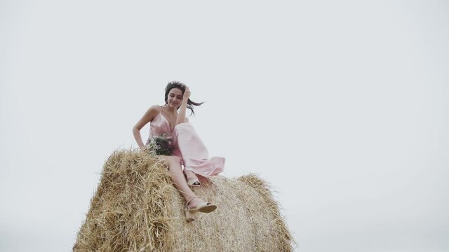 Attractive, happy lady sits in pose on large haystack in field, wind blows dress