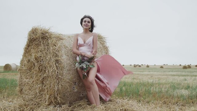 Seductive view of elegant lady poses at haystack in field during strong wind