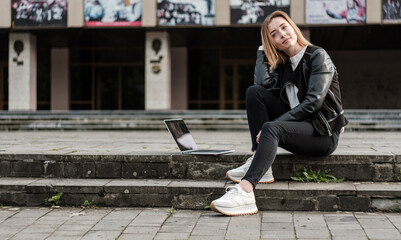 Blonde girl is resting after working on a laptop on the steps in the city.