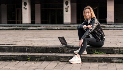 Blonde girl is resting after working on a laptop on the steps in the city.