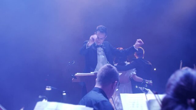 the male conductor waves his hands for the orchestra in smoke and colored light on the stage, and on the front stage the vocalists stand embracing and bowing