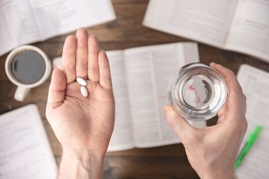 Work stress concept. Hands holding pills and glass of water above Workspace desk with pharmaceutical pills, books, pen, paper, and cup of coffee