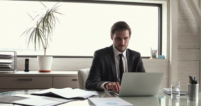 Successful satisfied young male company director wearing suit relaxing at workplace taking break after finishing computer work, chilling on chair, dreaming, feels peace of mind puts hands behind head.