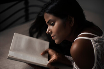 A young and attractive dark skinned Indian Bengali woman in lingerie reading book in a casual mood in white background. Indian lifestyle and Boudoir photography.