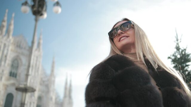 A Young Stylish Blonde Woman In Sunglasses In Fur Coat Poses Against The Background Of Famous Monuments Duomo In Milan. Bright Sunny Day