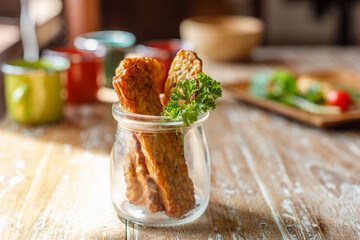Fried tempeh or tempe (traditional Indonesian soy product) in a small glass jar on wooden surface. Side view. With space. Vertical image.