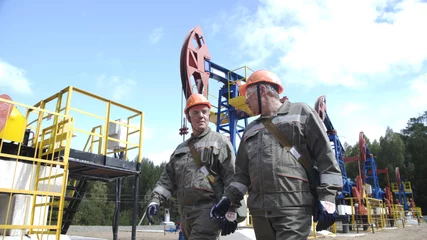 Tuinposter Two male oil workers walking and talking near oil pump jacks. Oil engineers overseeing drilling rig of crude oil production. Pumping crude oil for fossil fuel energy with drilling rig in oil field. © spocktv