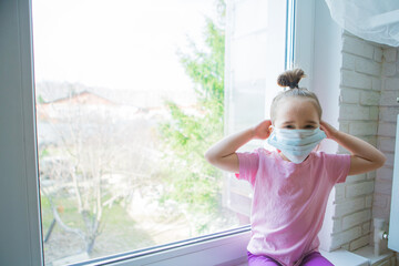 the girl in the medical mask leaned against the window. the girl looked out the window. a person is bored in self-isolation. I look at the world through glass. sad face