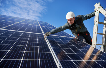 Man electrician standing on ladder and installing photovoltaic solar panels under blue cloudy sky. Worker wearing safety helmet and gloves. Concept of alternative energy, power sustainable resources.