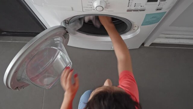 A girl loads dirty laundry into a washing machine while sitting on the floor in an apartment. Laundry day, housework