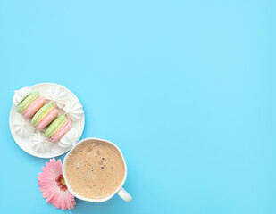 The cup of cappuccino and delicious macarons with white merengues on white plate  on blue background with pink flower. Happy day, breakfast concepts. Greeting card. Flat lay style with copy space.
