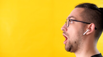 Very surprised man with glasses and headphones looks to the left side of the screen. Profile portrait of an attractive adult unshaven man on a yellow background. Emotional guy opened his mouth