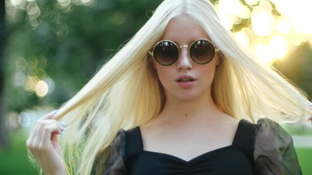 Cute young caucasian blonde girl in round stylish sunglasses and a black dress effectively fluffs her long hair with her hands with manicure against a background of trees and grass of a city park.
