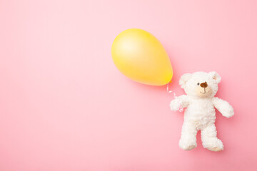 Smiling white teddy bear holding one yellow balloon on light pink table background. Pastel color....