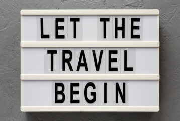 'Let the travel begin' on a lightbox on a gray surface, top view. Flat lay, from above, overhead. Close-up.