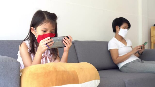 asian mother and daughter are wearing face mask, playing smartphone, and keeping social distancing while stay at home. covid-19 or coronavirus, new normal lifestyle concept